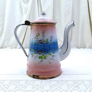 Peut inclure: Une cafetière en émail rose et blanche vintage avec un motif floral bleu et or. La cafetière a une poignée et un bec.