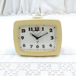 May include: A cream-colored alarm clock with black numbers and hands. The clock face reads "DISCRETO" and "JAZ".