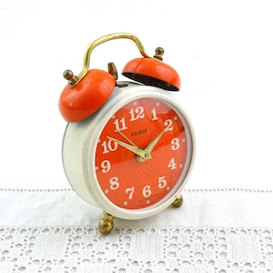 Puede incluir: Despertador Kaiser vintage con cuerpo blanco, esfera y campanas naranjas y detalles dorados. La esfera muestra la palabra "Kaiser" y agujas doradas. El reloj tiene un diseño clásico con una estética retro.
