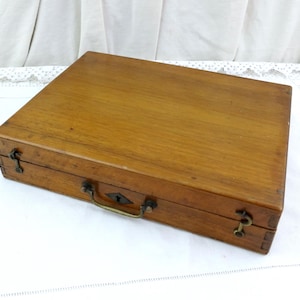 May include: A rectangular wooden storage case with a smooth, medium-brown finish. The box features a hinged lid, a central metal handle, a keyhole escutcheon, and two metal latches on the front side to secure the contents within the wooden frame.