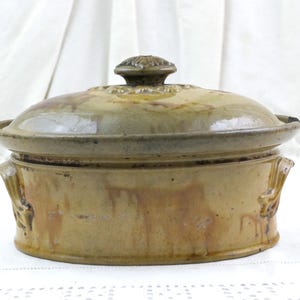 May include: An oval-shaped ceramic casserole dish with a matching lid, featuring a mottled tan and brown glaze. The vessel includes two side handles and a decorative knob on the lid, showcasing a glossy finish with visible color variations.