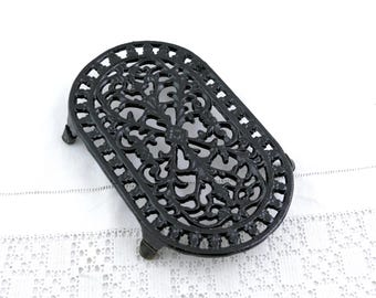 May include: Black cast iron trivet, oval in shape, featuring an intricate floral pattern. The trivet has four small feet and is designed to protect surfaces from hot items. A decorative kitchen accessory.