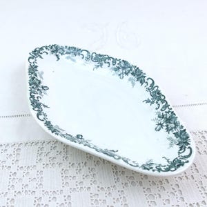 May include: An elongated, diamond-shaped white ceramic dish featuring a dark green floral and scrollwork border pattern along the rim. The dish has a smooth, glossy finish and a shallow basin, showcasing intricate botanical details in the print.