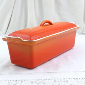 Puede incluir: Un molde para pan rectangular de cerámica naranja con tapa y asa a juego. El molde tiene un borde blanco alrededor de los bordes. El molde está sobre una superficie blanca.