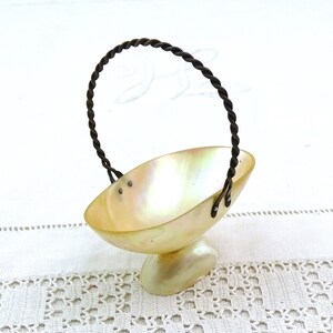 May include: A small, white, iridescent shell basket with a twisted wire handle. The basket is on a white crocheted surface.