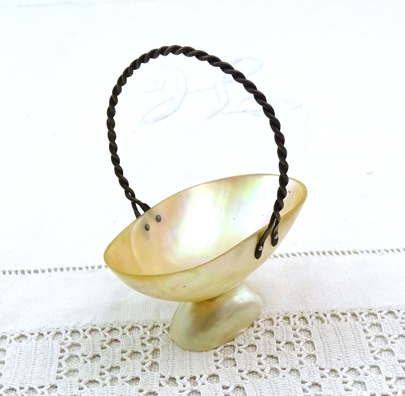 May include: A small, white, iridescent shell basket with a twisted wire handle. The basket is on a white crocheted surface.