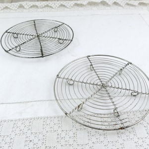 May include: Two round, vintage metal cooling racks. Each rack features concentric circular wires connected by radial wires, creating a grid pattern. The racks have small feet and are set against a white background.