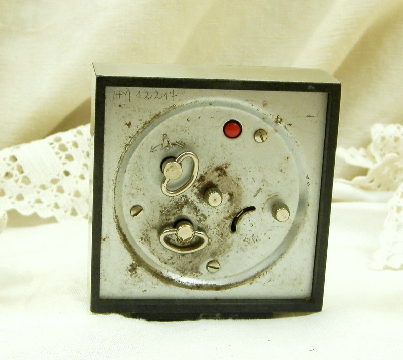 Puede incluir: Un reloj despertador vintage plateado con una esfera negra y un bot&oacute;n rojo. El reloj tiene una inscripci&oacute;n manuscrita en la parte posterior que dice "HPM 122214".