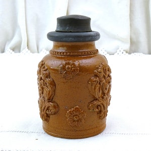 May include: A brown ceramic jar with a metal lid and intricate floral designs. The jar has a slightly rounded shape and a textured surface.