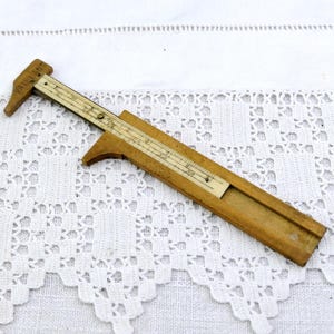 Può includere: Un calibro in legno vintage con una scala scorrevole e un piccolo braccio di misurazione. Il calibro è realizzato in legno chiaro con una scala color avorio a contrasto. Lo strumento è progettato per misurare piccoli oggetti. Lunghezza di circa 15 cm.