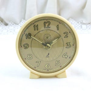 Può includere: Una sveglia analogica rotonda color crema con quadrante dorato. Il quadrante presenta numeri arabi neri, un piccolo sotto-quadrante e un logo a forma di uccello sopra la scritta JAZ e MADE IN FRANCE. L'orologio ha due lancette metalliche e un supporto.