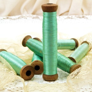 Op de afbeelding: Zes spoelen met mintgroene draad, gewikkeld op houten spoelen, liggen op een wit kant tafelkleed. De spoelen zijn allemaal even groot en hebben een vergelijkbaar ontwerp.
