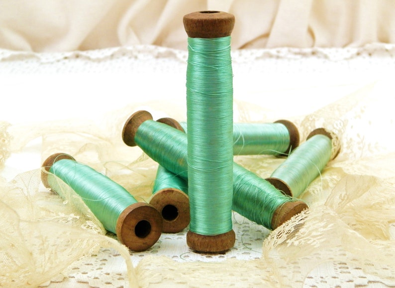 Op de afbeelding: Zes spoelen met mintgroene draad, gewikkeld op houten spoelen, liggen op een wit kant tafelkleed. De spoelen zijn allemaal even groot en hebben een vergelijkbaar ontwerp.