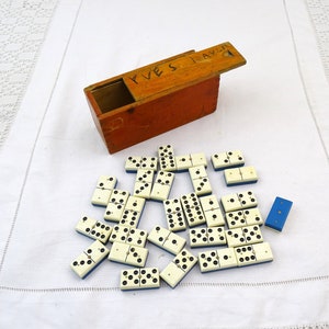 May include: A vintage wooden domino box with the text "FUES TRAVEL" on the lid. The box is open and contains a set of white dominoes with blue edges. The dominoes are scattered on a white tablecloth.