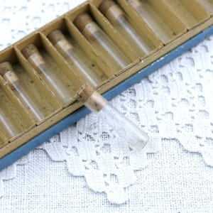 May include: A vintage set of small glass vials with cork stoppers, arranged in a divided cardboard box. The box has a blue border and the vials are clear, showcasing their contents. The background is a white lace fabric.