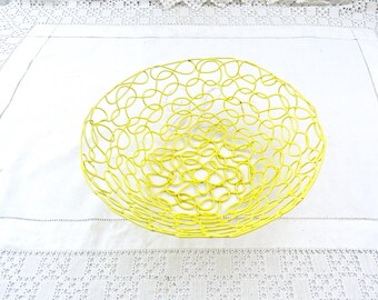 Può includere: Un cestino in filo metallico giallo brillante di forma arrotondata. Il cestino è realizzato con anelli di filo metallico interconnessi, creando un design aperto e arioso. Il cestino è su una superficie bianca con un bordo in pizzo.