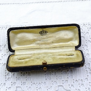 Op de afbeelding: Een open, antieke zwarte sieradendoos met een goudkleurig interieur. De doos is rechthoekig met afgeronde hoeken en een kleine gouden sluiting. De binnenkant is gevoerd met een vervaagde, crèmekleurige stof. Een klein kroonembleem is zichtbaar in de deksel.