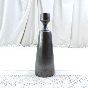 Puede incluir: Un portavelas alto de metal gris oscuro con una base acampanada y una parte superior cilíndrica. El portavelas tiene una superficie lisa, ligeramente texturizada y un diseño minimalista. Se muestra sobre un mantel de encaje blanco.