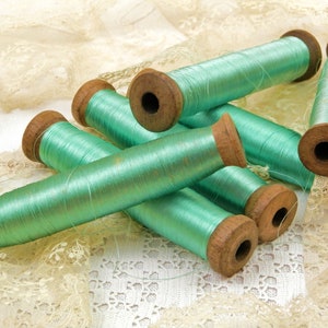Op de afbeelding: Zes spoelen tealgroen garen, elk met een houten spoel. De spoelen liggen op een witte kant achtergrond.