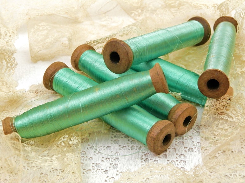 Op de afbeelding: Zes spoelen tealgroen garen, elk met een houten spoel. De spoelen liggen op een witte kant achtergrond.