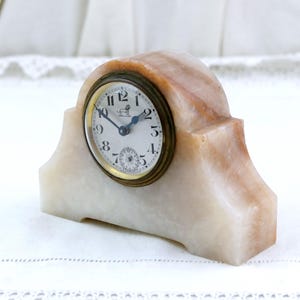 May include: A vintage marble clock with a cream and peach-colored stone base. The clock face is white with black numerals and hands, and a small seconds dial. The clock has a brass-colored bezel and a decorative lion emblem.