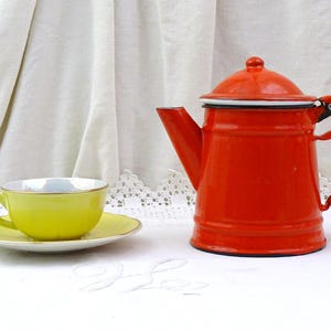 Può includere: Una teiera di smalto rosso con un bordo bianco e una tazza da tè e un piattino gialli su una tovaglia bianca.