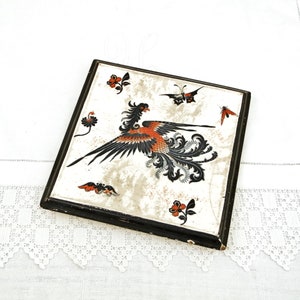 Puede incluir: Un azulejo de cerámica con marco negro, fondo blanco y diseño de fénix negro y naranja. El azulejo también presenta una mariposa y detalles florales.
