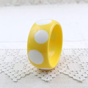 May include: A yellow bangle bracelet with large white polka dots. The bracelet is made of a glossy material and is set against a white lace background. The bracelet is a bold, eye-catching accessory.
