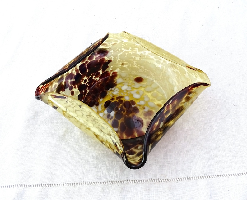 May include: A square-shaped glass bowl with a mottled brown and yellow pattern. The bowl has a wavy edge and is made of clear glass with brown and white flecks.