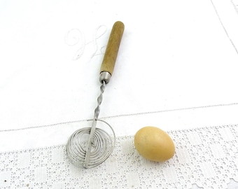 Può includere: Un separatore di uova vintage con manico in legno e frusta a spirale. Il separatore di uova è in metallo e si trova accanto a un uovo marrone chiaro. Gli oggetti sono su una superficie bianca strutturata.