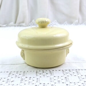 May include: A small, pale yellow lidded container with a round shape. The lid has a small knob on top. The container has a decorative design on the sides. The background is a white, textured surface.