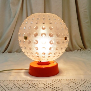 May include: A white globe lamp with a textured surface and an orange base. The lamp is turned on and illuminated.