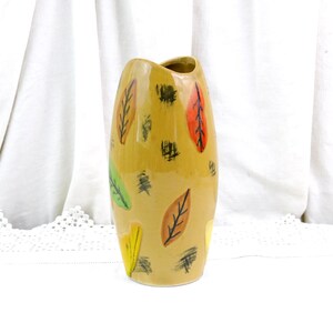 May include: A tall, ceramic vase with a mustard-yellow base. The vase is decorated with colorful leaf designs in shades of red, green, brown, and yellow. The vase has a unique, curved shape and is suitable for displaying flowers.