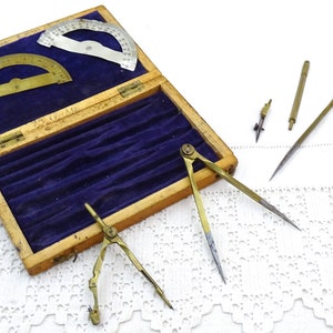 Puede incluir: Una caja de madera con un interior de terciopelo azul que contiene un juego de cuatro compases de latón antiguo y dos transportadores. La caja está abierta y los compases están dispuestos sobre una superficie de crochet blanco.