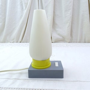 Peut inclure: Une lampe de table en céramique blanche avec une base jaune et un interrupteur gris. La lampe a un design côtelé et est posée sur une nappe blanche.