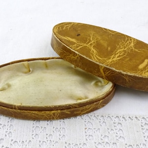 Può includere: Una scatola ovale in similpelle marrone con un interno in raso bianco. La scatola è aperta e il coperchio è appoggiato sul lato.