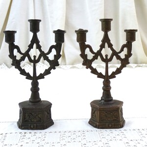 May include: Two antique bronze candle holders with a decorative, ornate design. Each holder has three candle arms and a hexagonal base.