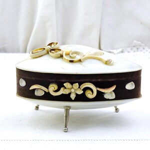May include: An oval jewellery box with a white lid and a dark brown base. The lid is decorated with shell-like embellishments. The base features decorative floral and shell accents. The box rests on three silver-coloured legs.