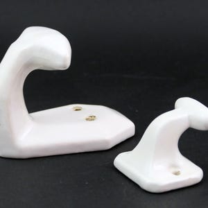Op de afbeelding: Twee witte keramische wandhaken met een glanzende afwerking. De grotere haak heeft een gebogen, langwerpig ontwerp met twee montagegaten in de basis, terwijl de kleinere haak een korter, T-vormig profiel heeft met één montagegat in de basis.