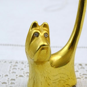 May include: Gold-colored metal ring holder shaped like a dog with rhinestone eyes. The dog is sitting with its tail curled up.