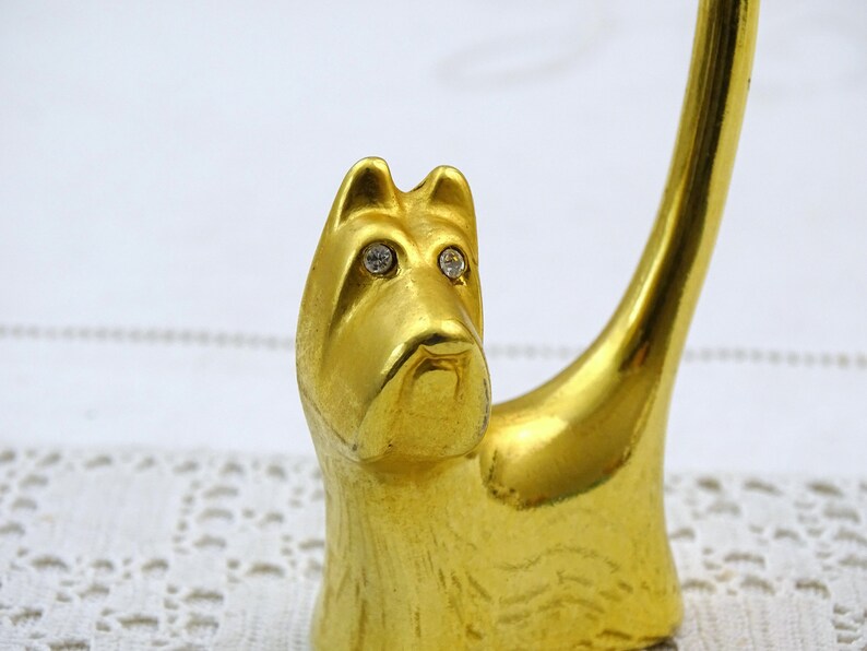 May include: Gold-colored metal ring holder shaped like a dog with rhinestone eyes. The dog is sitting with its tail curled up.