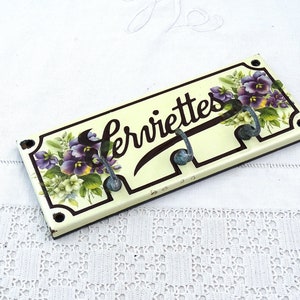 May include: A white ceramic towel rack with three hooks and a floral design. The rack is labeled "Serviettes" in black lettering.