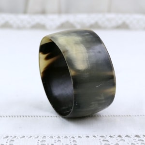 Puede incluir: Un brazalete circular ancho elaborado con cuerno natural pulido. La superficie presenta un acabado liso y semitranslúcido con un patrón jaspeado en tonos gris oscuro, negro y beige crema, mostrando variaciones orgánicas en el color.