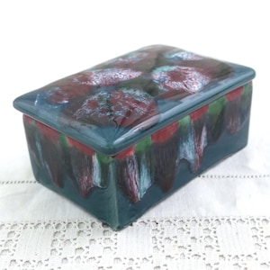 May include: Rectangular ceramic box with a lid. The box is teal with a glossy finish and features a design of red and green abstract shapes on the lid. The sides have a dripped paint effect in shades of red, green, and teal.