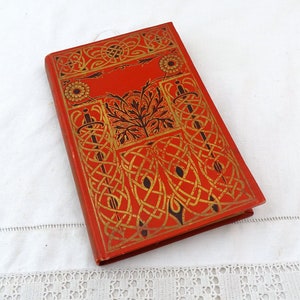 Op de afbeelding: Een rood boek met een gouden en zwart Keltisch knoopontwerp op de omslag. Het boek heeft een decoratieve rand en een bloemmotief in het midden.