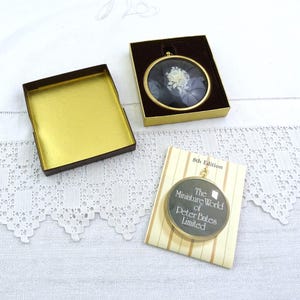 May include: An open brown box with a gold interior sits next to a gold-framed, circular pendant with a white floral design on a dark background. A second pendant with text, "The Miniature World of Peter Bates Limited," rests on a striped card.
