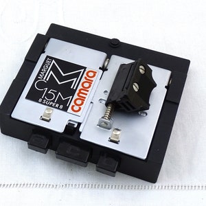 May include: A black plastic Super 8 film cartridge loader with a silver metal plate. The cartridge is labeled "Marguet C15M 8 Super 8" and "camara".