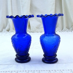 May include: Two cobalt blue glass vases with a ruffled top and a floral design. The vases are on a white lace tablecloth.