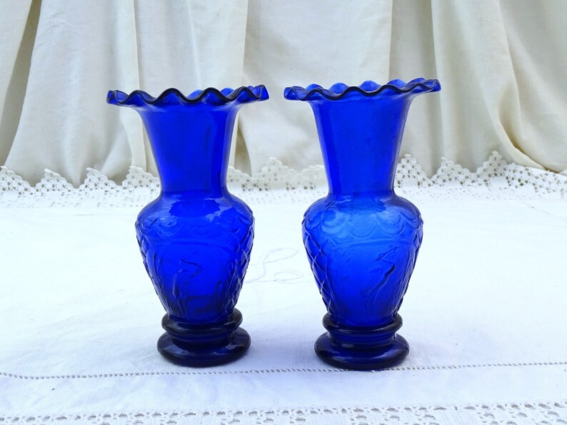 May include: Two cobalt blue glass vases with a ruffled top and a floral design. The vases are on a white lace tablecloth.