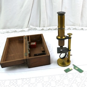 May include: A vintage brass microscope with a circular base, a stage, and an adjustable mirror, positioned next to an open wooden storage box containing a small red container and internal compartments, with two green textured pieces nearby.